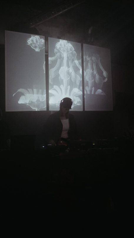 DJ hinter einem Mischpult mit projizierten Licht- und Grafikeffekten im Hintergrund.