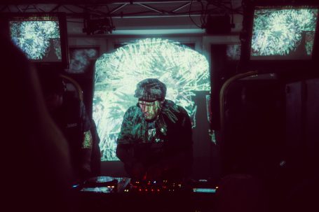 Ein DJ steht vor Bildschirmen mit bunten visuellen Effekten während eines Auftritts.