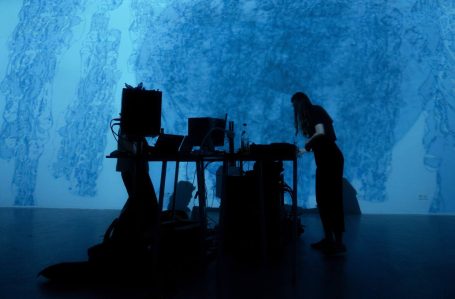 Person steht an einem Tisch, umgeben von Schatten und blauer Beleuchtung.