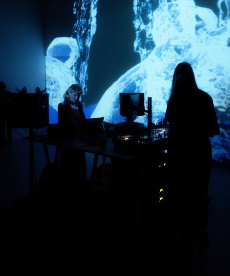 Zwei Personen an einem Tisch, mit Projektionen und elektronischer Ausrüstung im Hintergrund.